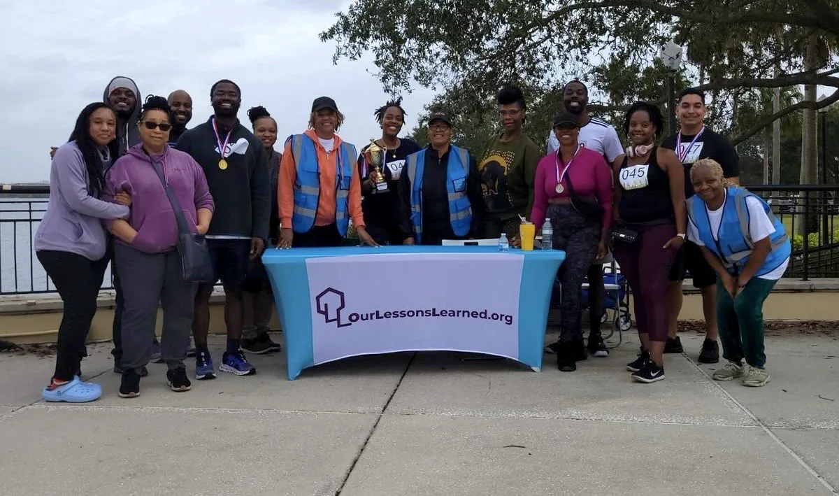 The Our Lessons Learned team and volunteers gathered together at a community walk in Orlando, behind an OurLessonsLearned.org banner