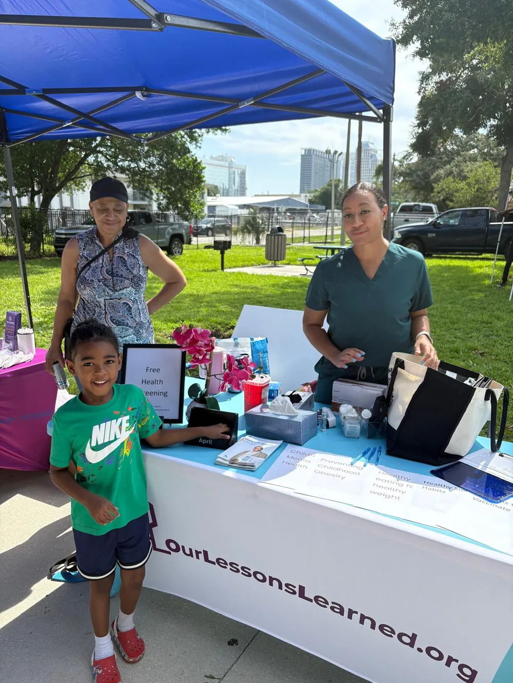 A nurse in scrubs at the free health screening table with a grandmother and a young boy
